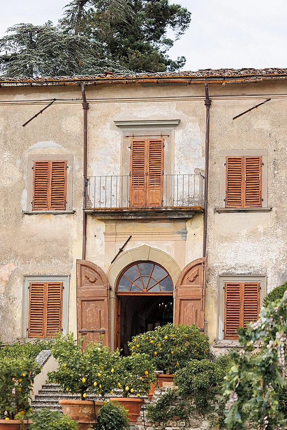 Villa entrance with rustic villa facade, wooden double doors and wood shutters, flanked by terracotta pots and citrus trees on stone steps