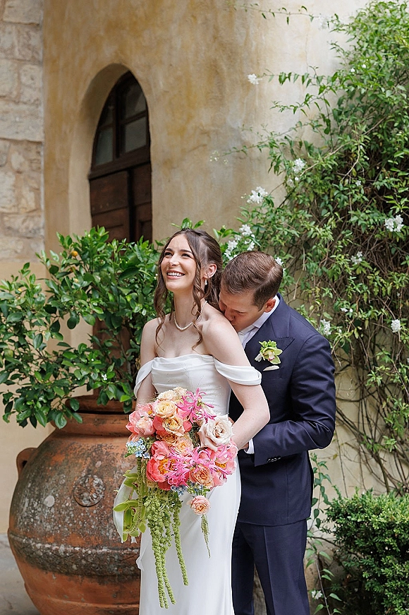 Couple portrait of groom kissing bride’s shoulder as she holds a colorful bouquet by a stucco wall and arched doorway with vines