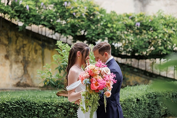 Couple portrait of bride and groom close embrace, touching foreheads as she holds a colorful bouquet by an ivy stone wall in a garden
