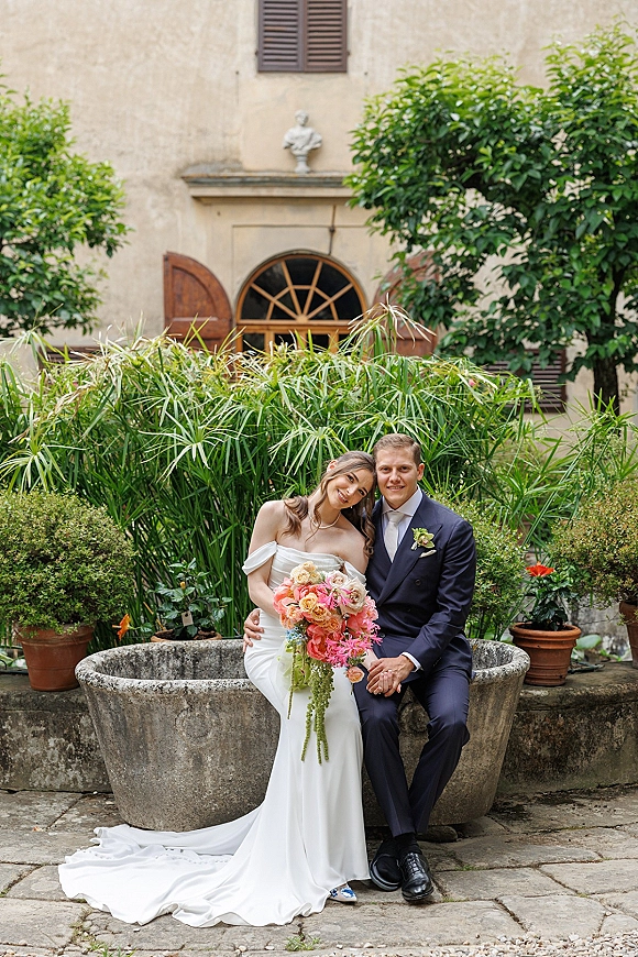Couple portrait of bride and groom seated, her colorful bridal bouquet in hand, in a courtyard garden with arched shuttered window.