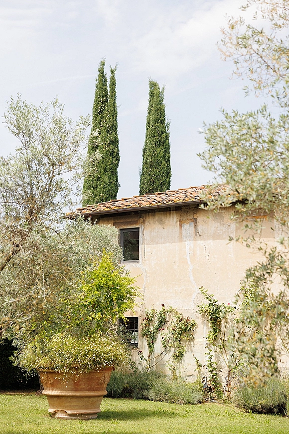 Villa exterior with rustic villa facade, terracotta roof tiles and flowering vines, framed by greenery, lawn, and blue sky