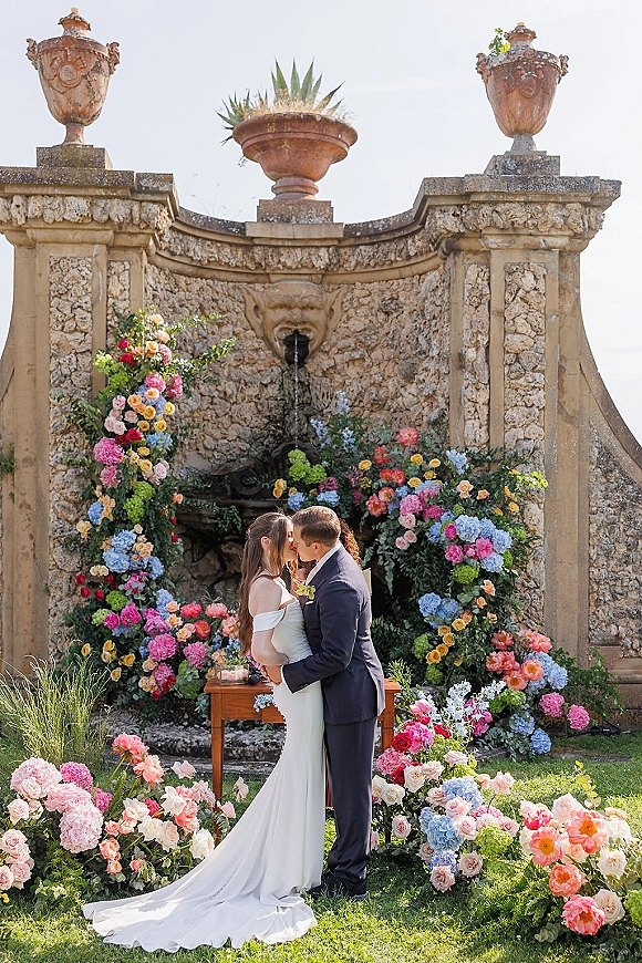 Wedding kiss portrait of bride and groom kissing beneath a rose and hydrangea arch, with a fountain and stone wall in the garden behind