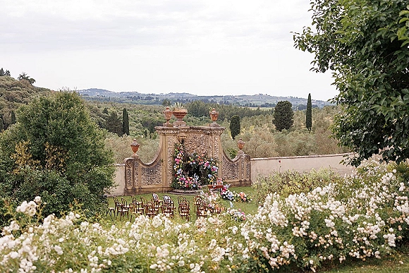 Outdoor ceremony setup with a floral ceremony arch, wood chairs, and ground florals against a stone wall on a garden lawn under cloudy skies
