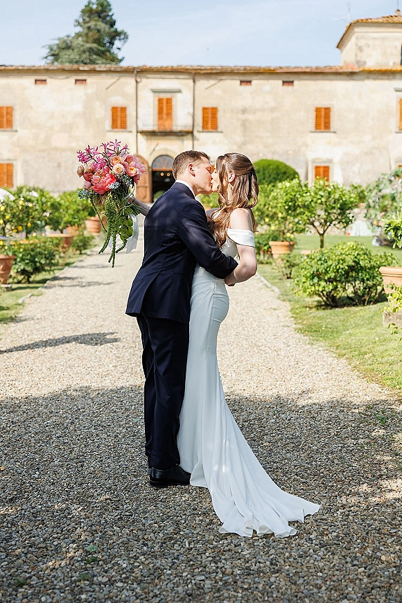 Wedding kiss portrait of bride and groom kissing on a garden path by a villa, bride holding a pink and coral bouquet in an off-shoulder dress