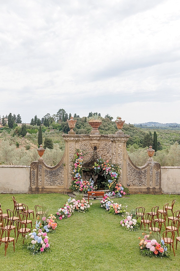 Ceremony setup for an outdoor wedding ceremony with floral aisle markers, wooden chairs, and an arch by a fountain and stone wall