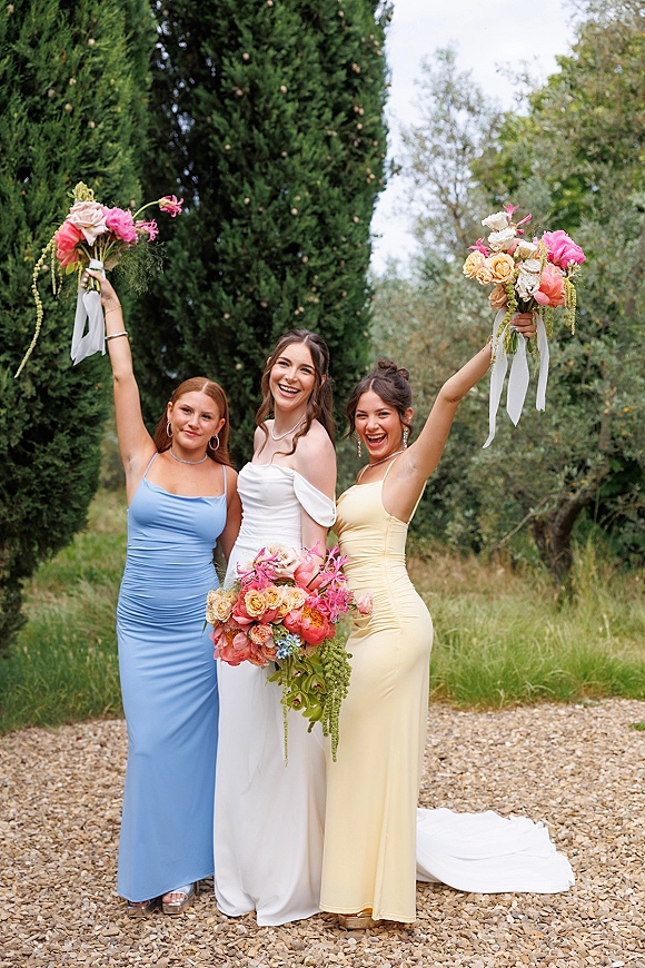Bridesmaid portrait with bridesmaid bouquet pose, smiling women raising colorful bouquets with ribbon streamers on a gravel path by evergreens