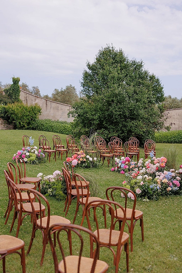 Ceremony setup with wood bistro chairs and garden ceremony seating, lined with pastel roses, peonies and hydrangea on a grassy lawn by a stone wall