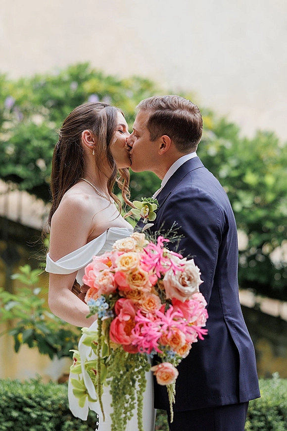 Wedding kiss as bride and groom kiss in side profile, her off-the-shoulder gown and bouquet with ribbon against vine-covered wall