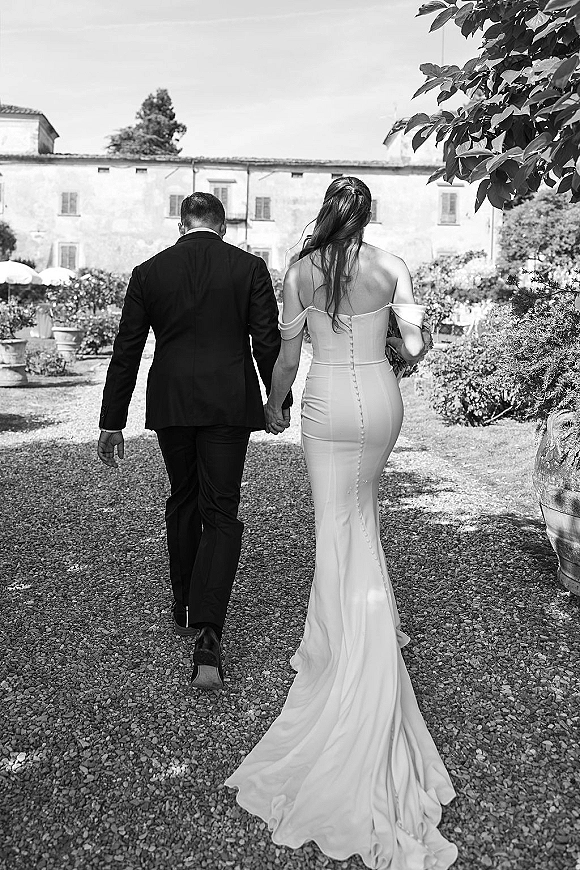 Couple portrait of newlyweds holding hands from behind, bride in off-shoulder gown with bouquet beside groom in tux on a garden gravel path