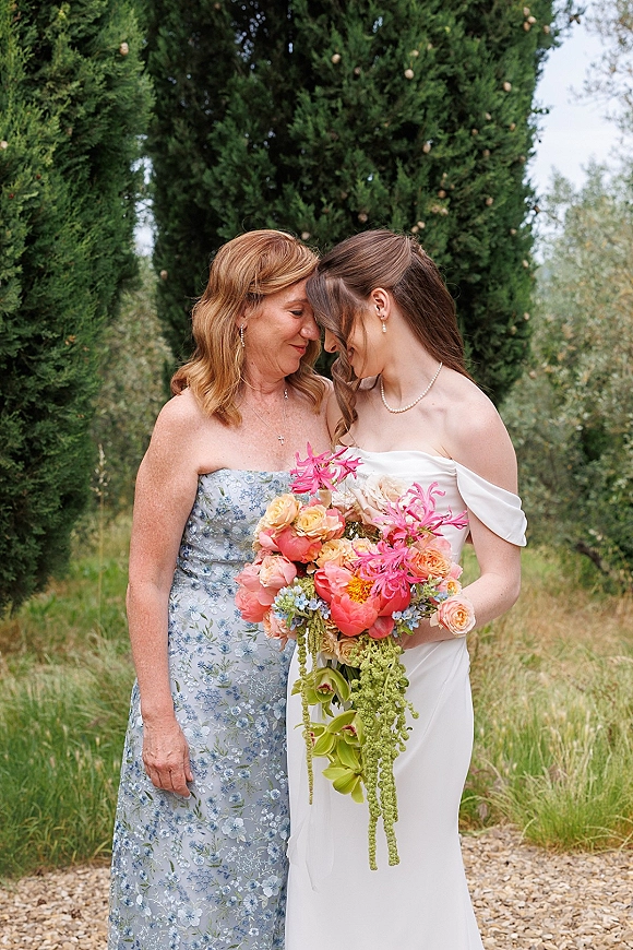 Bride portrait holding a cascading bouquet of pink and coral flowers with orchids, wearing an off-the-shoulder dress in a meadow by evergreens