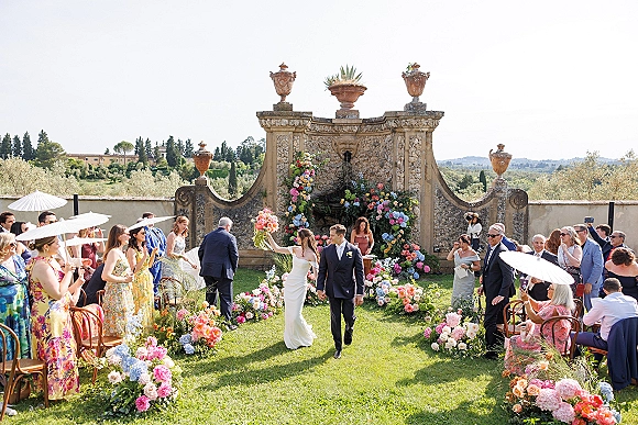 Wedding recessional as bride and groom walk the aisle holding hands, bride bouquet in hand beneath a floral arch by a stone fountain