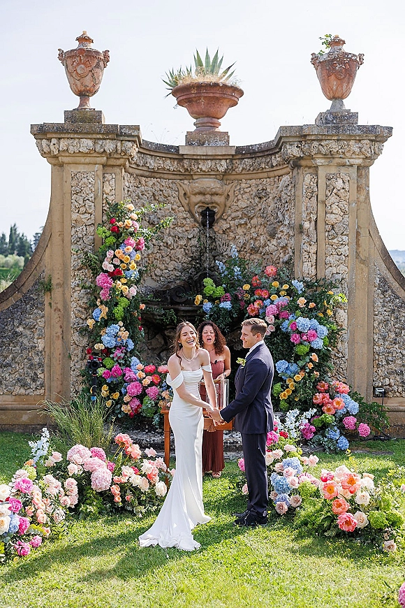 Wedding vows at an outdoor wedding ceremony as bride and groom hold hands with officiant under a floral arch by a stone fountain backdrop