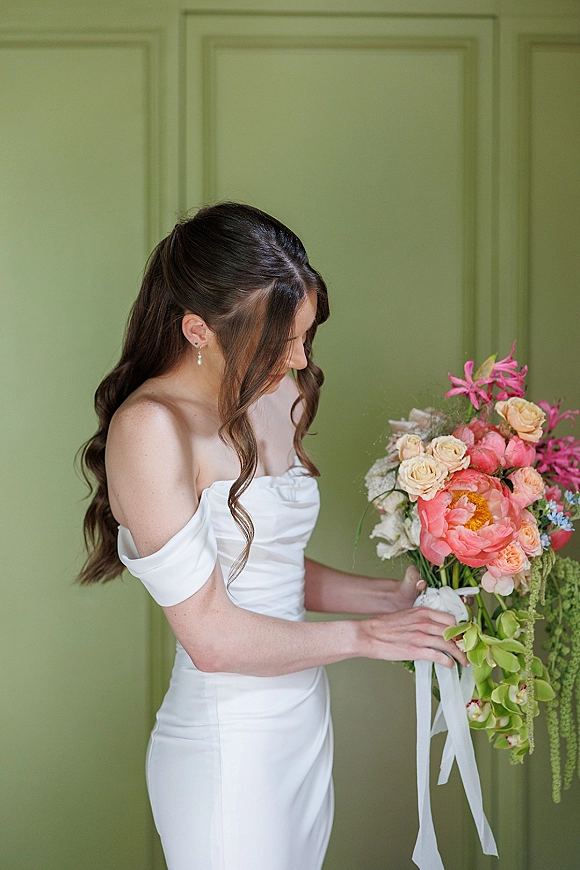 Bridal portrait of a bride holding bouquet in an off the shoulder wedding dress, long ribbon trailing, posed against a green paneled wall