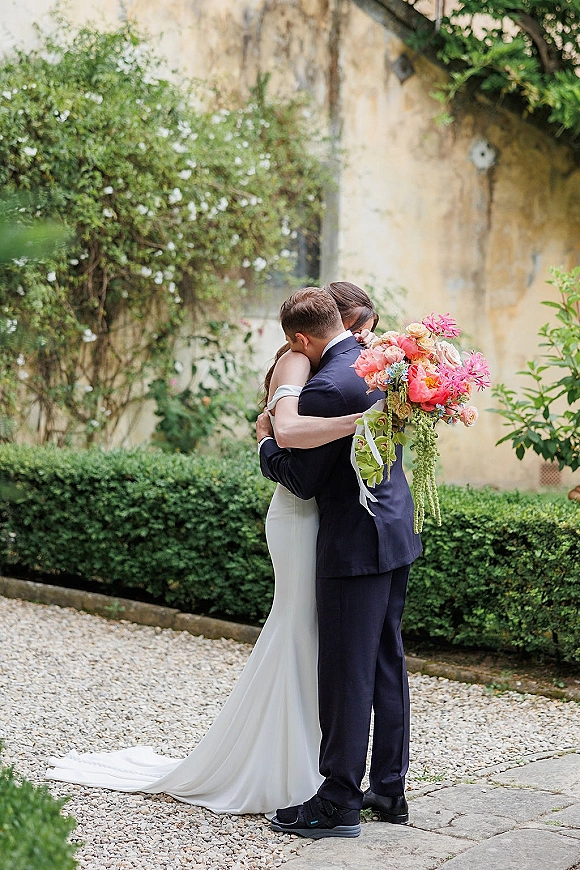 Wedding couple portrait with bride and groom hugging, holding a pink and coral bouquet with trailing greenery on a garden gravel path, stone wall behind