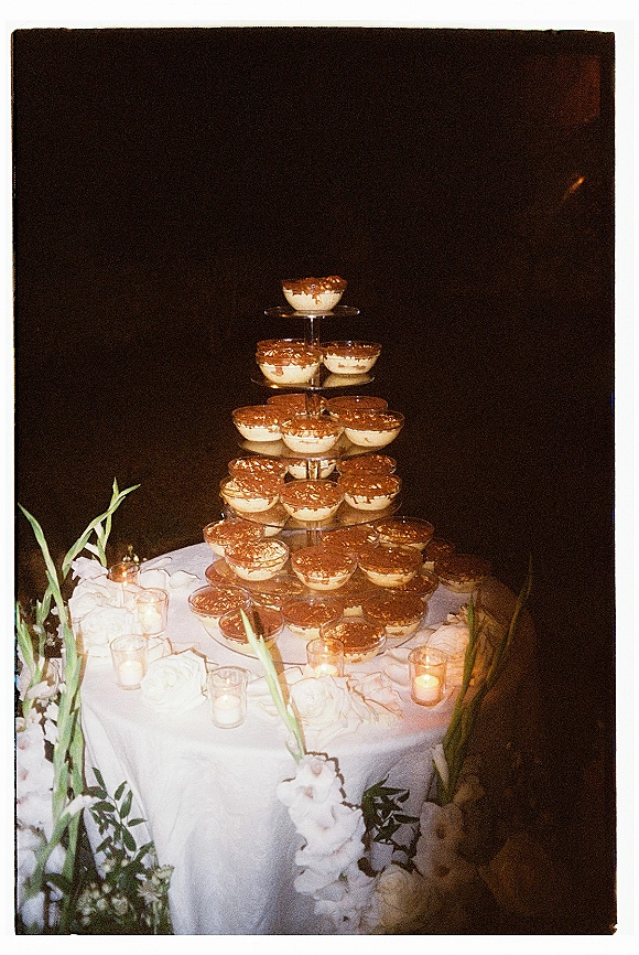 Dessert table with tiered dessert display of tiramisu cups in glass cups, cocoa dusting, and votive candles on a white tablecloth against a dark backdrop