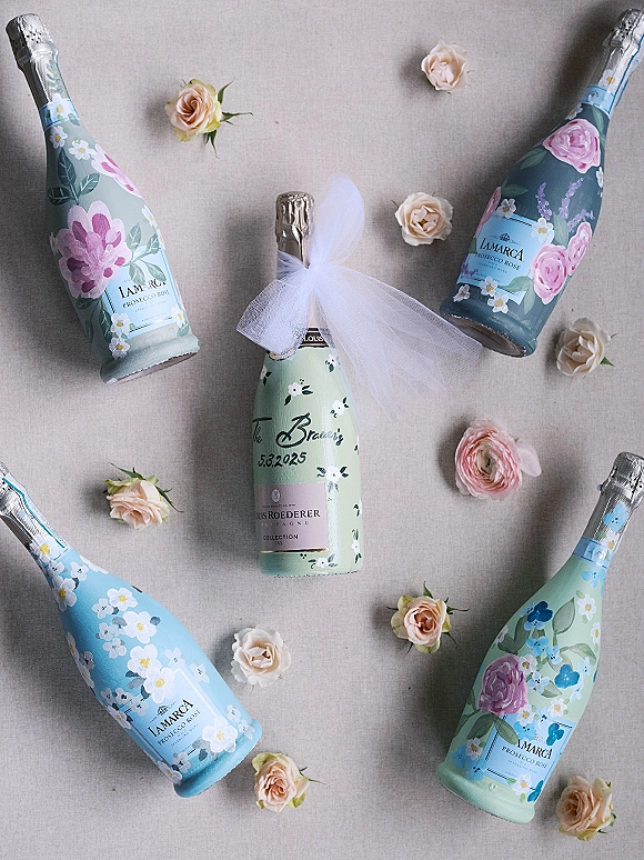 Champagne bottles flatlay of wedding champagne bottles with floral labels, a tulle bow, and rose accents on a linen fabric backdrop