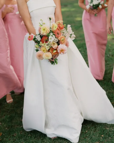 Bridal bouquet with dahlia wedding bouquet blooms in peach and pink, held against a strapless wedding dress on a grass lawn