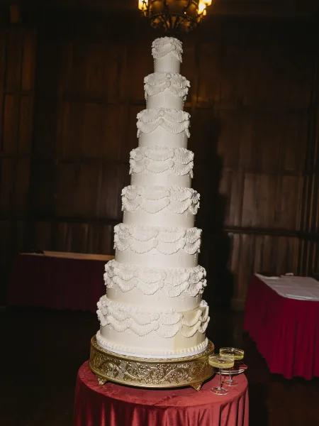 Wedding cake, a multi tier wedding cake displayed on a gold cake stand against wood paneling beneath a chandelier in an indoor venue.