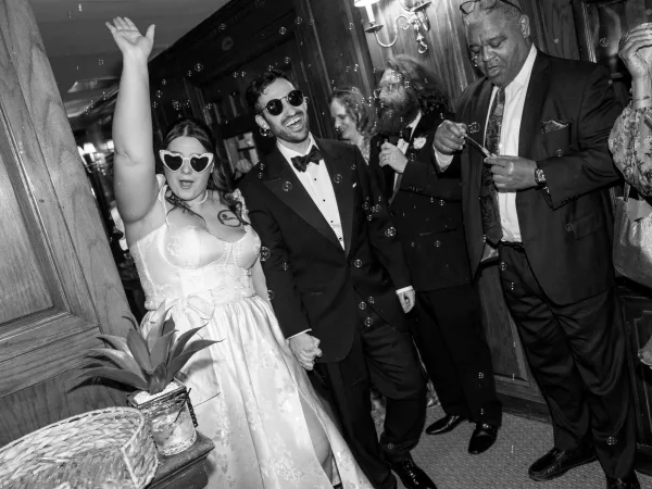 Wedding exit during a bubble send off as the newlyweds walk hand in hand, the bride in heart sunglasses, in a wood-paneled hallway.