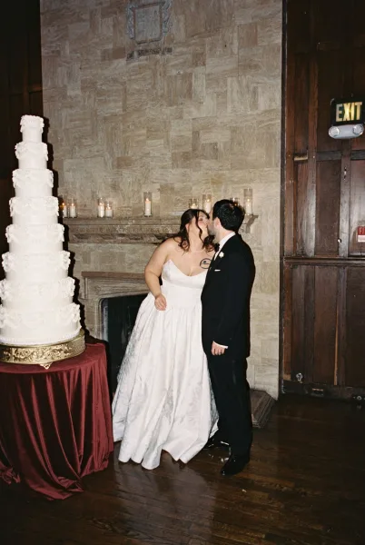 Wedding kiss portrait capturing a reception kiss moment beside a wedding cake, set before a stone fireplace and wood paneling.