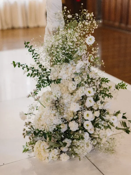 Ceremony flowers line the aisle in elegant aisle flower arrangements with white roses, set on wood flooring before sheer curtains.