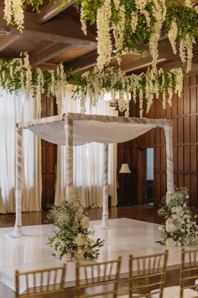 Wedding chuppah showcasing elegant ceremony canopy decor with hanging wisteria, set indoors against wood-paneled walls and beams.
