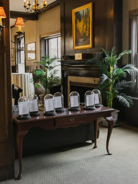 Seating chart display arranged as a glass cloche seating chart with black and white cards, set before a fireplace mantel and dark wood paneling.