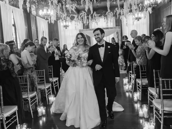 Wedding recessional with a bride holding bouquet as she and her groom walk a candlelit aisle in a wood-paneled hall.