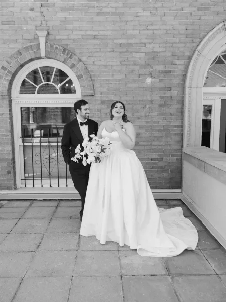 Couple portrait of the bride and groom laughing, her gown’s long train sweeping the stone terrace against a brick wall with arched windows.