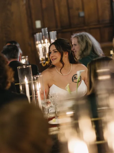 Bride portrait, bride at reception seated by hurricane candles against warm wood paneling, gazing softly as guests chat at the table.