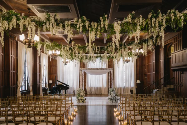 Ceremony setup for an indoor wedding altar with hanging florals, framed by wood-paneled walls and tall windows over a dark wood floor.