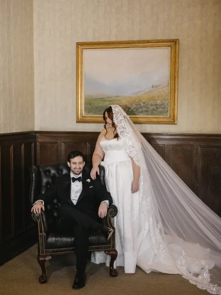 Couple portrait of a bride standing and groom seated, her lace veil draping as they pose in a wood-paneled room with a framed painting.