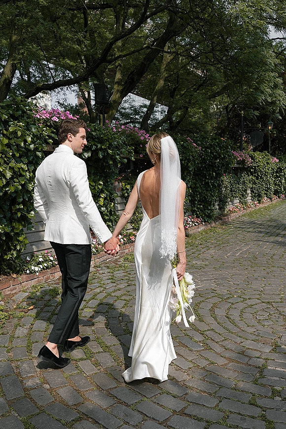 Couple portrait of bride and groom walking away hand in hand, cathedral veil and bouquet trailing on a cobblestone garden path with ivy wall