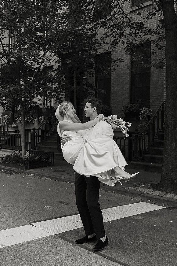Couple portrait of groom carrying bride in a bridal veil, laughing on a city street crosswalk with brick townhouse steps behind them