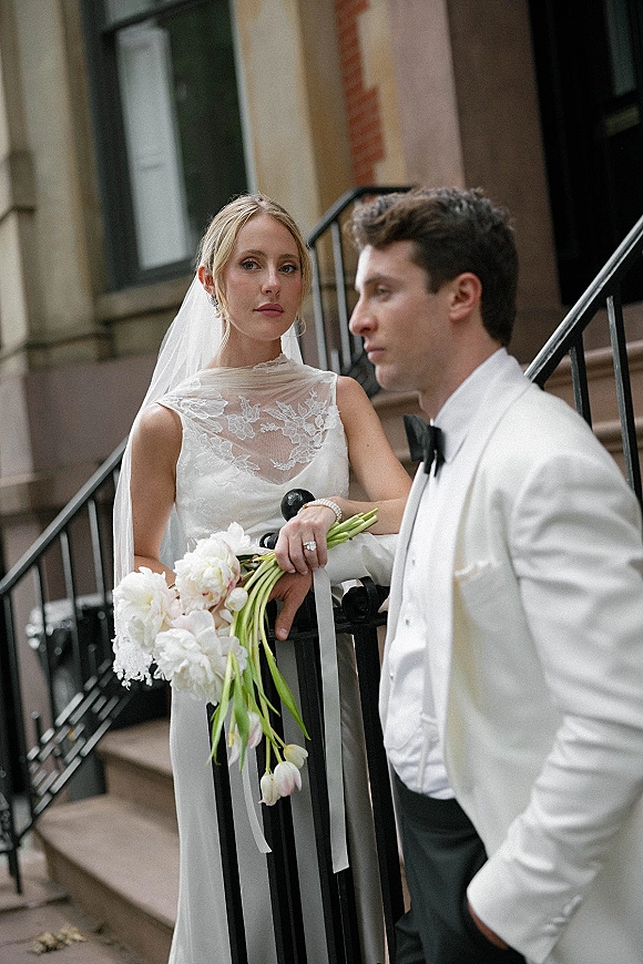 Couple portrait of bride in lace veil holding a ribbon bouquet with groom in white tuxedo, posed on brownstone steps by wrought iron railing