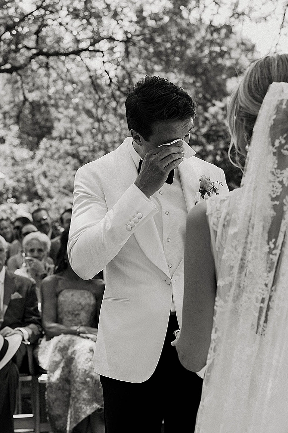 Ceremony moment as groom cries at altar in white tuxedo jacket and black bow tie, bride in lace veil, guests seated among trees behind