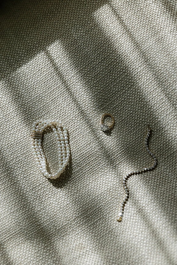 Wedding jewelry flatlay featuring a diamond engagement ring with pearl and tennis bracelets on textured fabric in natural light shadows