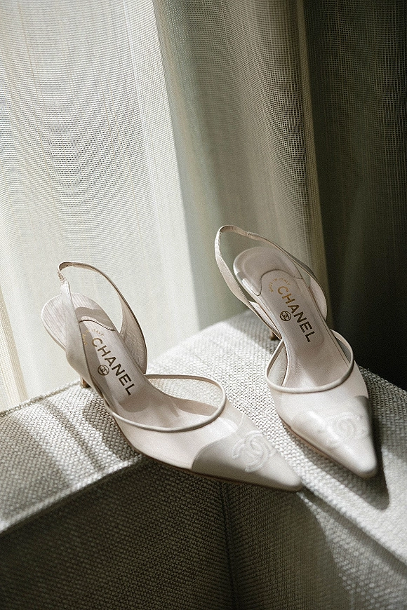 Bridal shoes, white slingback heels with pointed toes and logo detail, resting on an upholstered ottoman by sheer curtains in window light