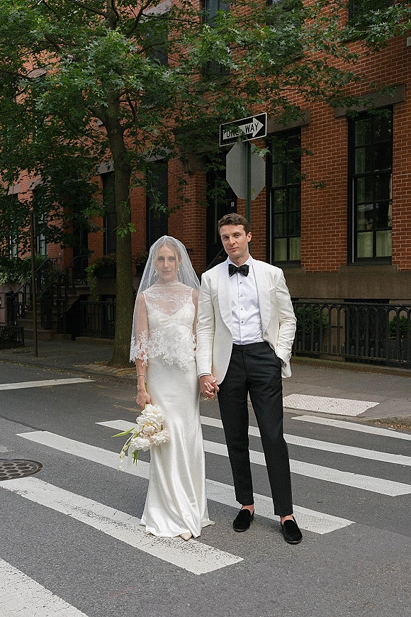 Couple portrait of bride and groom holding hands on a city crosswalk, her lace veil and bouquet beside his white dinner jacket and boutonniere