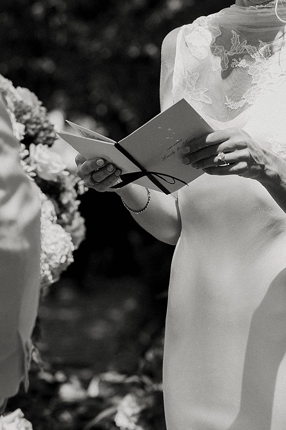 Wedding vows as bride reading vows from a vow book with ribbon, hands showing ring and bracelet, lace high-neck dress amid greenery