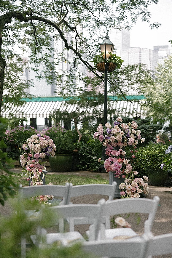 Ceremony floral arch with asymmetrical floral arch clusters of roses, hydrangeas, and orchids on pedestals, beside white chairs in a garden setting