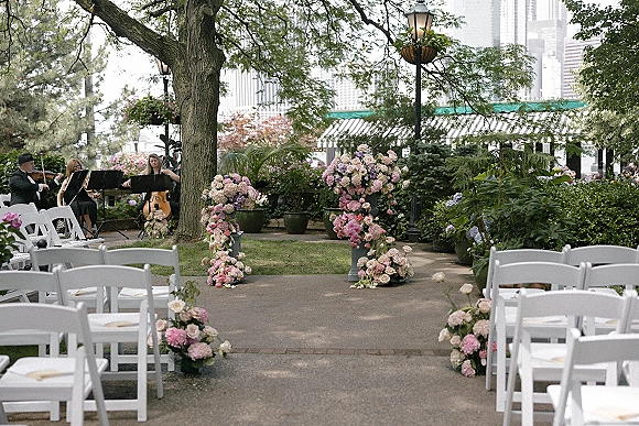 Ceremony aisle decor with pink hydrangea and rose arrangements on pedestals beside white folding chairs along a garden path, one lantern nearby