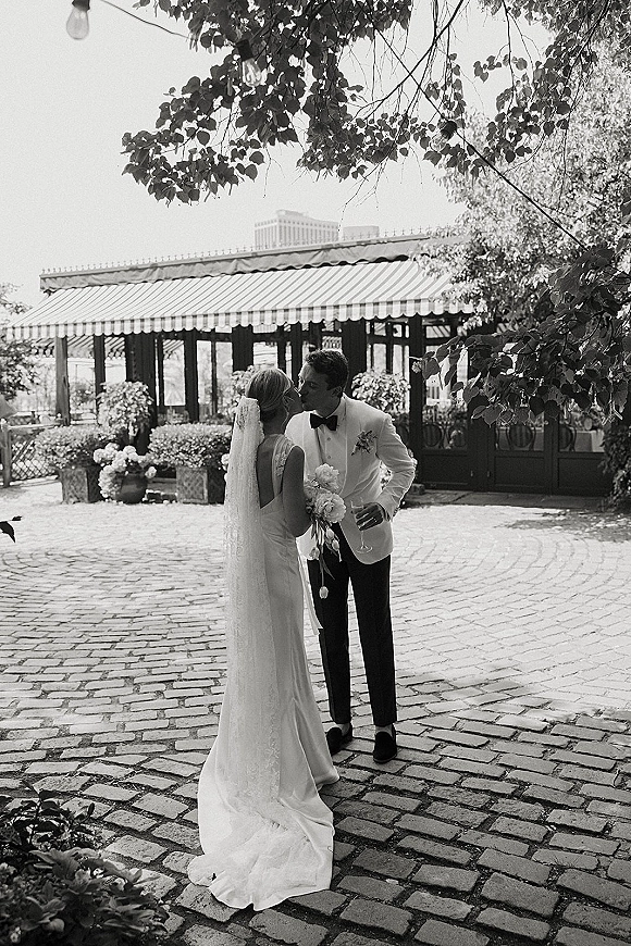 Wedding couple portrait of bride and groom kissing under string lights in a cobblestone courtyard, her long veil flowing as he holds a wine glass