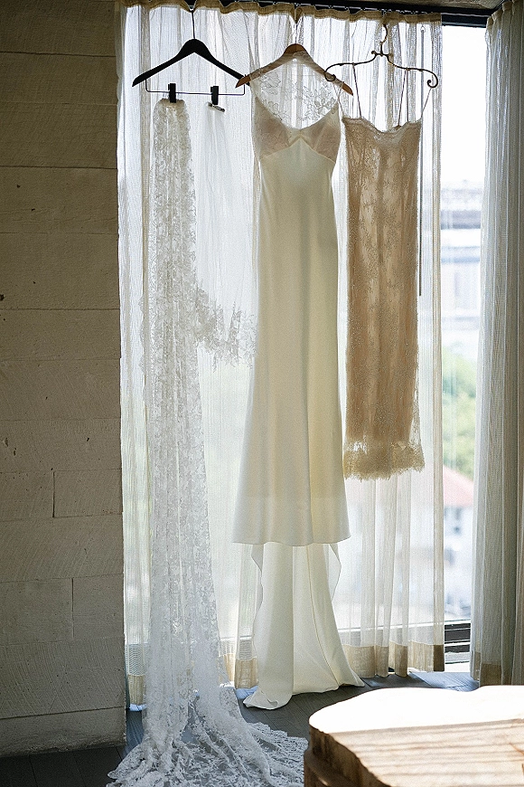 Wedding dress hanging by the window with a lace veil accent, softly lit by window light against a wood plank wall