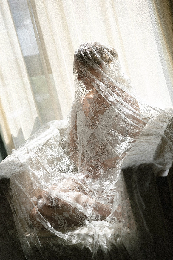 Bridal portrait of a bride with veil in soft window light, lace veil over her face as she sits by curtains in an upholstered chair
