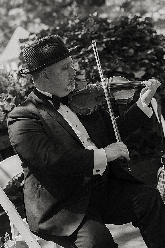 Wedding violinist, ceremony violinist in tuxedo and fedora plays with bow beside a white folding chair amid dappled garden light.