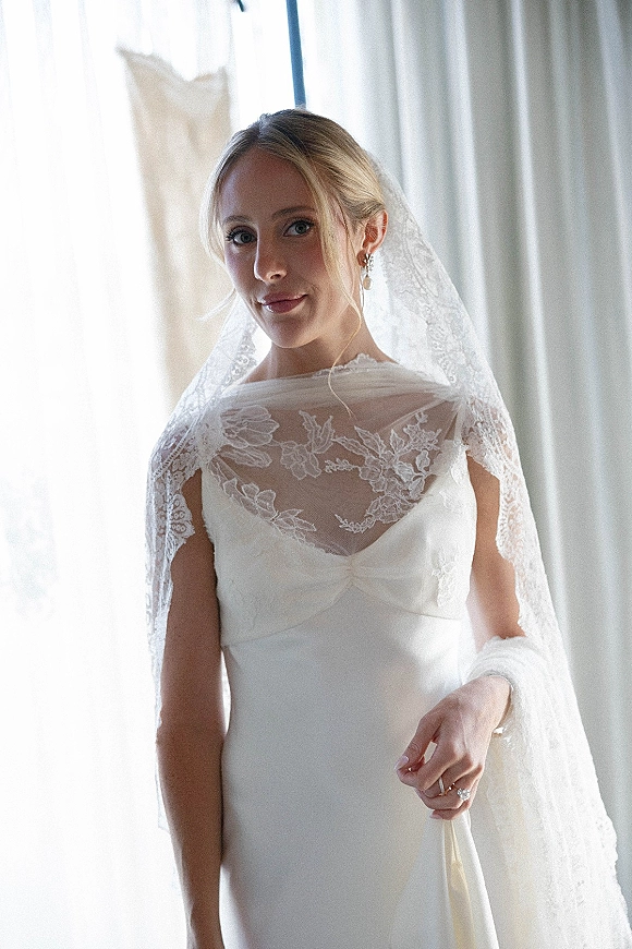 Bridal portrait of a bride in a white wedding dress and lace veil, pearl drop earrings and ring glowing by window light with sheer curtains