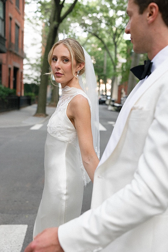 Couple portrait of bride looking back while holding hands with groom, her long veil flowing on a tree-lined city crosswalk with brick buildings