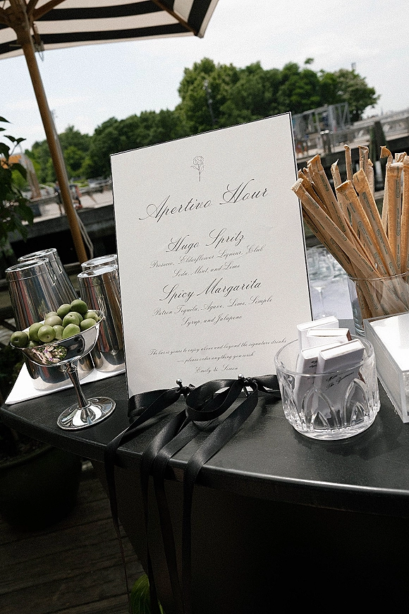 Wedding bar menu calligraphy sign tied with a black ribbon beside cocktail glasses, olives, and straws on a waterfront deck