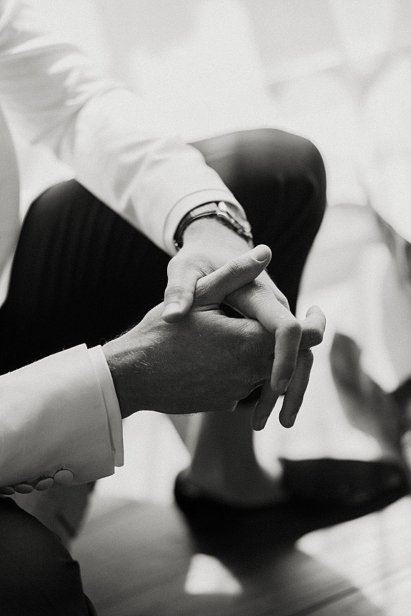Wedding hands close-up of bride and groom holding hands, showing cufflinks and wristwatch against outdoor pavement with soft shadows